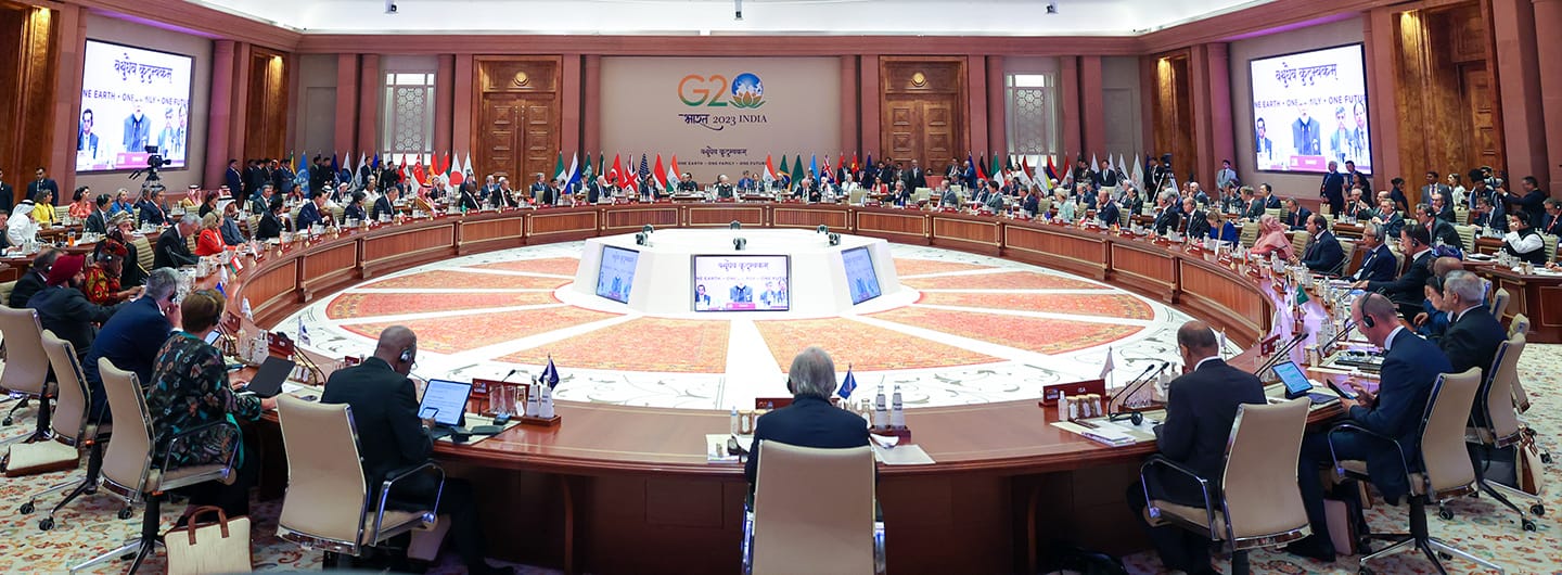 Large circular table with delegates, flags, and "G20 INDIA 2023" sign. Screens display "One Earth, One Family, One Future".